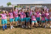 A sports team in pink shirts and turquoise shorts waving arms in the air and smiling